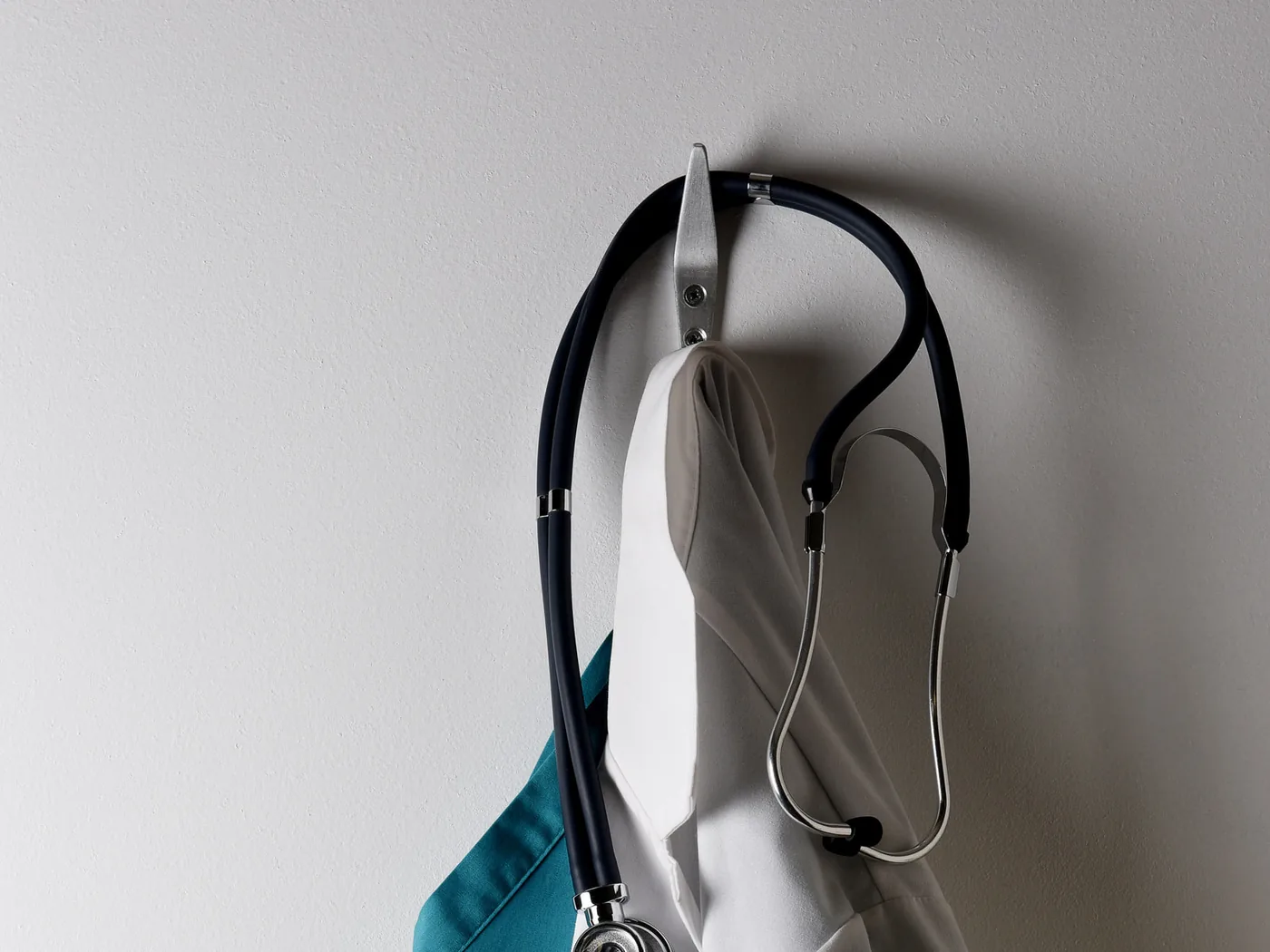 A white lab coat, a teal scrub top and a stethoscope hanging on a wall hook